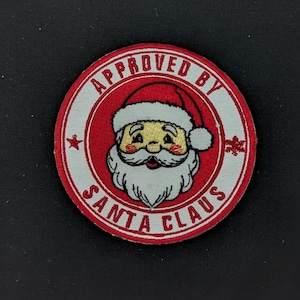 Puede incluir: Un parche redondo, rojo y blanco con el texto "APPROVED BY SANTA CLAUS" que rodea una imagen de dibujos animados de Papá Noel. La ilustración de Papá Noel presenta una barba blanca, mejillas rosadas y un gorro rojo.