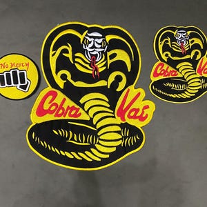 Könnte beinhalten: Gestickte Aufnäher mit dem Cobra Kai Logo und Text. Die Aufnäher zeigen ein großes Kobra-Kopf-Design mit den roten Worten "Cobra Kai" und einen kleineren Aufnäher mit einer Faust und den Worten "No Mercy".