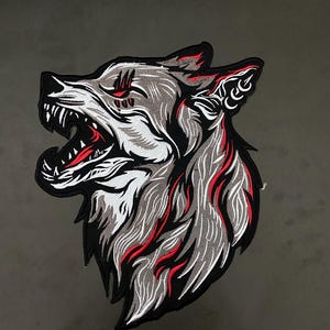 May include: Embroidered patch featuring a snarling wolf's head. The design uses shades of gray, white, and red against a black outline. The wolf's mouth is open, revealing sharp teeth. The patch is likely for jackets or bags.