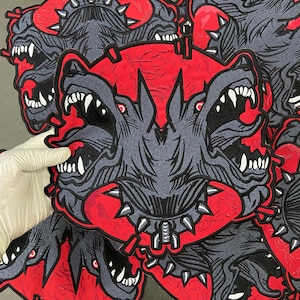 Può includere: Toppa ricamata con un disegno di cane a tre teste con denti affilati e occhi rossi. La toppa ha uno sfondo rosso e un cane grigio con accenti neri. Il cane ha un collare a punte.