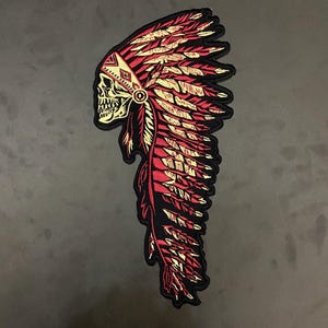 Könnte beinhalten: Gestickter Aufnäher mit einem Totenkopf, der einen indianischen Kopfschmuck trägt. Der Kopfschmuck ist rot und goldfarben, mit aufwendigen Federdetails. Der Schädel ist cremefarben und der Aufnäher hat einen schwarzen Rand.