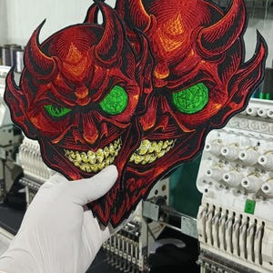 Puede incluir: Dos parches bordados con un diseño de cara de diablo. Los parches son principalmente rojos con contornos negros, ojos verdes y dientes amarillos. El diablo tiene cuernos y una sonrisa amenazante. Los parches se sostienen frente a una máquina de bordar.