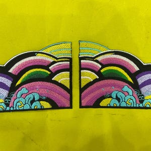 Op de afbeelding: Twee geborduurde patches met een kleurrijk ontwerp. Elke patch heeft een zwarte rand, met lagen roze, geel, groen en paars, en turquoise golven. De patches staan op een gele achtergrond.