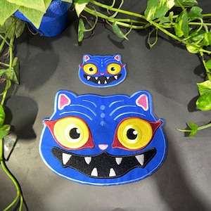 Könnte beinhalten: Zwei blaue, katzenförmige, gestickte Aufnäher. Der größere Aufnäher hat gelbe Augen, rote Augenbrauen und einen schwarzen Mund mit weißen Zähnen. Der kleinere Aufnäher spiegelt das Design wider. Aufnäher sind wahrscheinlich für Kleidung oder Accessoires.