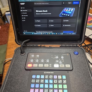 May include: A black Stream Deck device with multiple buttons and a screen displaying the Stream Deck website. The device is inside a black case. The screen shows the text "Stream Deck" and "Discover a world inside your device."