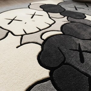 Striking modern rug in a minimalist living room. It features a dense wool texture and an abstract pattern of overlapping Kaws style companion figures with crossed eyes and gloved hands, rendered in rich shades of cream, light grey, dark grey, and black. The plush hand-carved design feels upscale and contemporary on a warm wood floor.