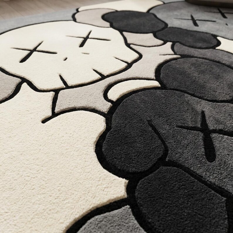 Striking modern rug in a minimalist living room. It features a dense wool texture and an abstract pattern of overlapping Kaws style companion figures with crossed eyes and gloved hands, rendered in rich shades of cream, light grey, dark grey, and black. The plush hand-carved design feels upscale and contemporary on a warm wood floor.