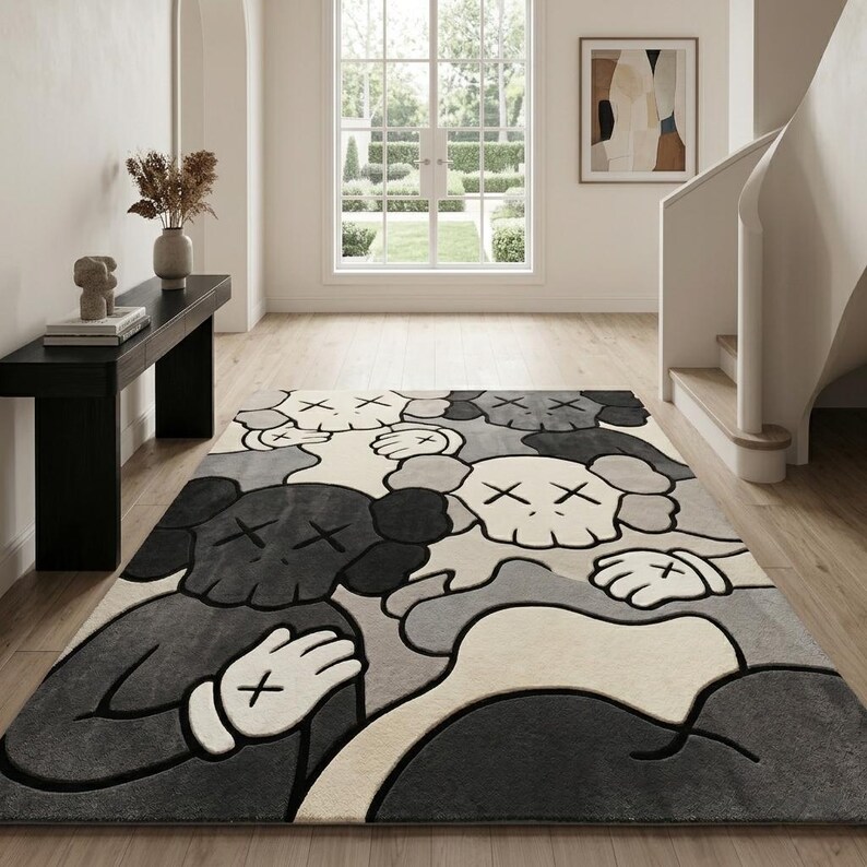 Striking modern rug in a minimalist living room. It features a dense wool texture and an abstract pattern of overlapping Kaws style companion figures with crossed eyes and gloved hands, rendered in rich shades of cream, light grey, dark grey, and black. The plush hand-carved design feels upscale and contemporary on a warm wood floor.
