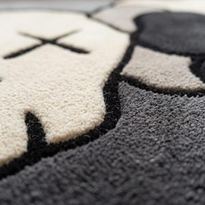Striking modern rug in a minimalist living room. It features a dense wool texture and an abstract pattern of overlapping Kaws style companion figures with crossed eyes and gloved hands, rendered in rich shades of cream, light grey, dark grey, and black. The plush hand-carved design feels upscale and contemporary on a warm wood floor.