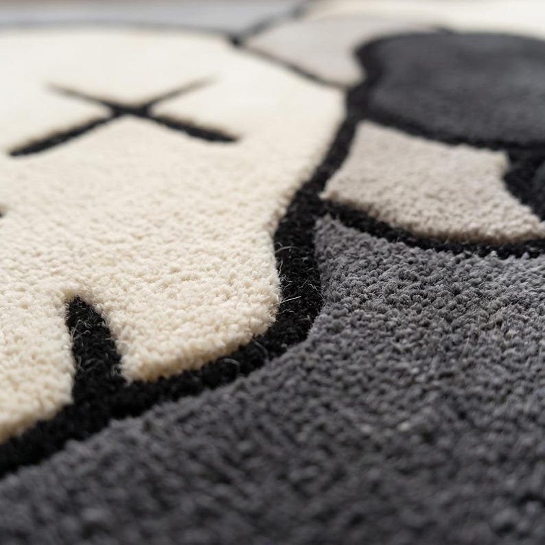 Striking modern rug in a minimalist living room. It features a dense wool texture and an abstract pattern of overlapping Kaws style companion figures with crossed eyes and gloved hands, rendered in rich shades of cream, light grey, dark grey, and black. The plush hand-carved design feels upscale and contemporary on a warm wood floor.