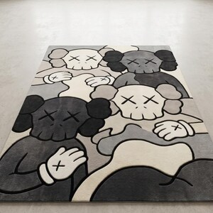 Striking modern rug in a minimalist living room. It features a dense wool texture and an abstract pattern of overlapping Kaws style companion figures with crossed eyes and gloved hands, rendered in rich shades of cream, light grey, dark grey, and black. The plush hand-carved design feels upscale and contemporary on a warm wood floor.