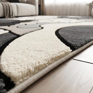 Striking modern rug in a minimalist living room. It features a dense wool texture and an abstract pattern of overlapping Kaws style companion figures with crossed eyes and gloved hands, rendered in rich shades of cream, light grey, dark grey, and black. The plush hand-carved design feels upscale and contemporary on a warm wood floor.