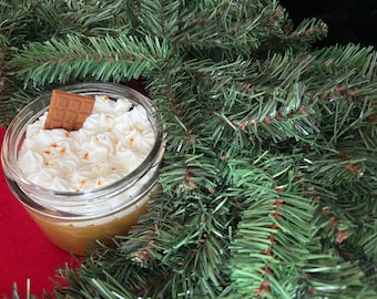 May include: A dessert in a clear glass jar, topped with white whipped cream and a piece of chocolate. The dessert sits in front of a backdrop of green pine needles, with a red surface visible below the jar.