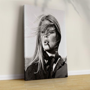 May include: Black and white portrait of a woman with long, flowing hair and a cigarette in her mouth. She is wearing a dark top and a scarf. The image is a canvas print, leaning against a wall.