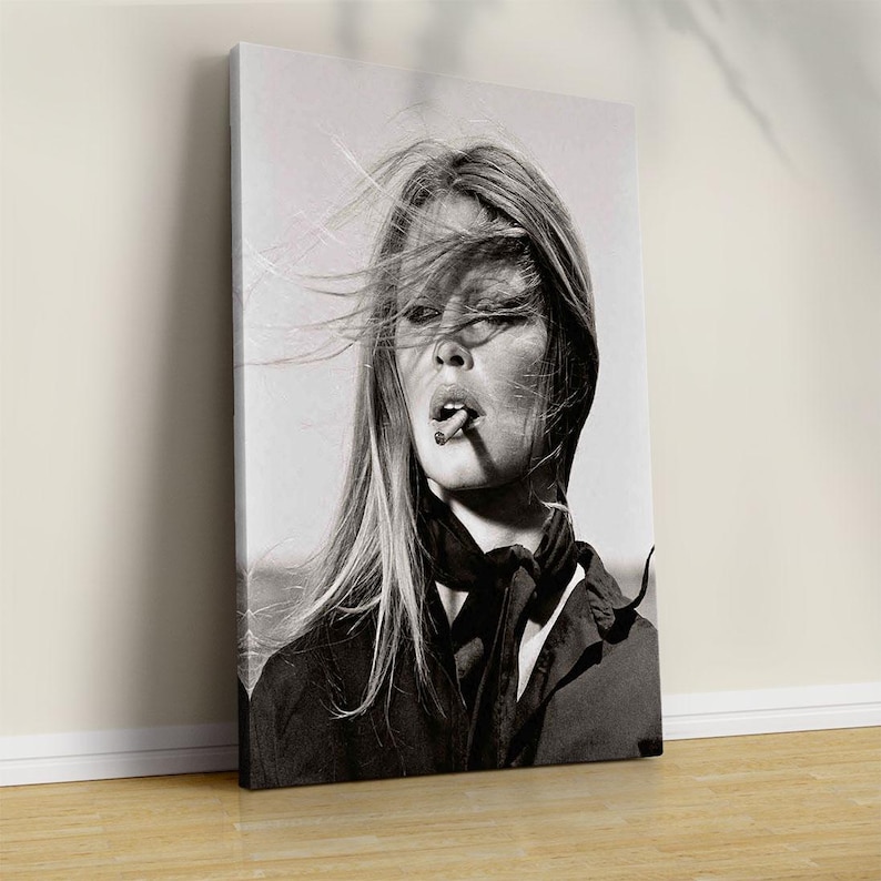 May include: Black and white portrait of a woman with long, flowing hair and a cigarette in her mouth. She is wearing a dark top and a scarf. The image is a canvas print, leaning against a wall.