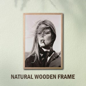 May include: Black and white framed print featuring a woman with wind-blown hair smoking a cigarette. The print is in a natural wooden frame. The text "NATURAL WOODEN FRAME" is printed at the bottom of the frame.