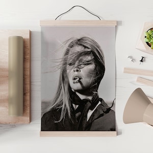 May include: Black and white print of a woman with long hair blowing in the wind, smoking a cigarette. The print is displayed on a wooden frame with a black hanging cord. The image is a portrait and is mounted on a white surface.