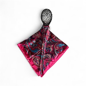 May include: A square, fuchsia-colored silk scarf with a paisley pattern in shades of red, blue, and gold. The scarf is displayed on a black head form, showcasing its design and texture. The scarf is likely a fashion accessory.