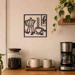 May include: A square black metal wall art piece featuring a cutout design of a moka pot, a steaming cup of coffee, scattered coffee beans, and a small potted plant. The minimalist line art is mounted on a plain wall above a wooden countertop.