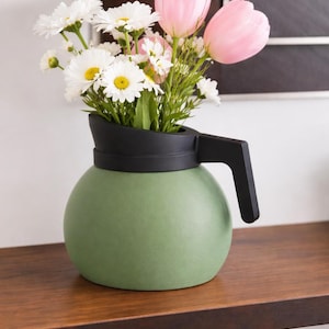 May include: A green ceramic vase shaped like a coffee pot, with a black handle and rim. The vase holds a bouquet of white daisies and pink tulips, creating a floral arrangement. The vase sits on a wooden surface.