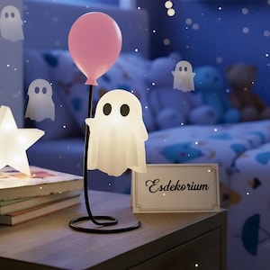 Puede incluir: Una lámpara de noche caprichosa con forma de fantasma sosteniendo un globo rosa, con una luz en forma de estrella y decoraciones de fantasmas flotantes. La escena está ambientada en un dormitorio infantil.