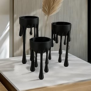 以下が含まれることがあります： ユニークな溶融デザインの黒い装飾プランター3つ。プランターはさまざまなサイズで、白と木のテーブルの上にあります。画像には、乾燥した植物とテキスト「3D Printable STL」も含まれています。