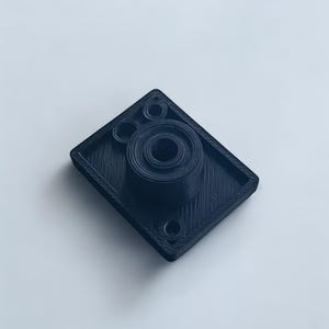 Puede incluir: Un componente negro rectangular impreso en 3D con una característica circular central y dos características circulares más pequeñas. El componente tiene una superficie texturizada y parece estar hecho de plástico. Dos pequeños agujeros son visibles en la base.