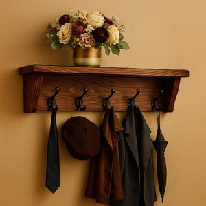 Può includere: Una mensola da parete in legno scuro con cinque ganci in metallo nero. Un vaso dorato riempito con rose color crema e rosso scuro poggia sulla mensola. Dai ganci pendono una cravatta blu scuro, un basco marrone, un cappotto marrone, un cappotto grigio e un ombrello.