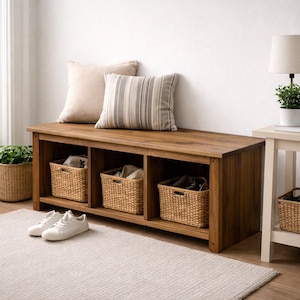 Può includere: Una panca contenitore rettangolare in legno con tre scomparti aperti, ciascuno contenente un cesto in vimini intrecciato. La panca è sormontata da un cuscino beige tinta unita e uno a righe beige e grigie. Un paio di scarpe da ginnastica bianche.