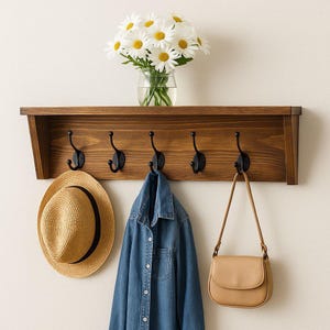 May include: A wooden wall-mounted coat rack with five black hooks. A straw hat, denim shirt, and tan purse hang from the hooks. A shelf above holds a glass jar of daisies. The wood is stained a warm brown.