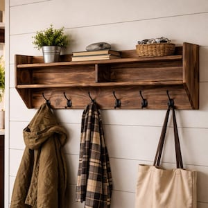 Peut inclure: Étagère murale en bois teinté foncé avec une étagère supérieure, une étagère inférieure et cinq crochets en métal noir. Elle contient une petite plante en pot, des livres, un panier avec des lunettes et des manteaux et un sac fourre-tout suspendus.