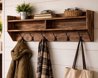 May include: A dark brown wooden wall-mounted shelf featuring a top storage ledge and six black metal hooks. The shelf holds a small potted green plant, two stacked books, and a woven basket. A brown jacket, a plaid scarf, and a beige tote bag hang below.