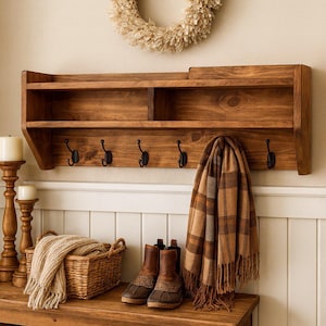May include: A dark stained wooden wall shelf with a top shelf, a lower shelf with five black hooks, and a small shelf on top. A brown and tan plaid scarf hangs from the hooks. A basket, boots, and candles are on the table below.