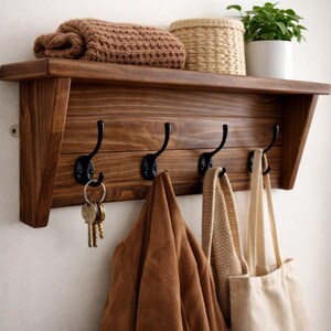 May include: A wooden wall-mounted shelf with four black metal hooks. A brown jacket, keys, a beige tote bag, and a knitted scarf hang from the hooks. A rolled brown blanket, a woven basket, and a potted plant sit on the shelf.