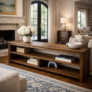 May include: A long, dark wood console table with three shelves. The top shelf holds a white vase of flowers, a woven basket, and a white bowl. The lower shelves display books and a white game controller. The table is in a living room.