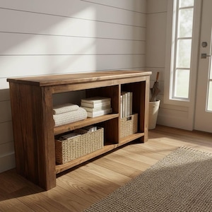 May include: A wooden storage console with open shelves and a flat top. The shelves hold folded towels, books, and woven baskets. The console is made of dark brown wood and sits on a light brown wooden floor.