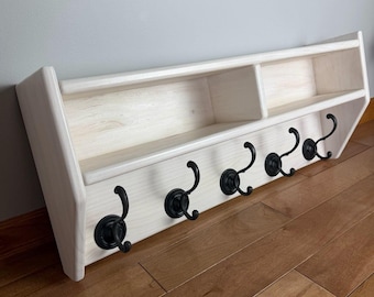 May include: A light-colored wooden wall shelf featuring two open storage compartments and five black metal hooks attached to the front panel. The shelf has a smooth finish with visible wood grain and angled side supports for mounting.