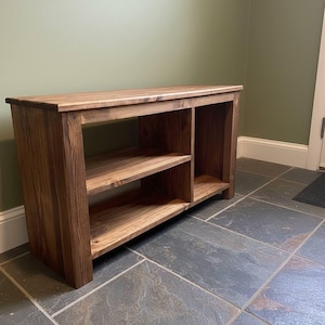 May include: A wooden console table with two shelves and a top surface. The table is made of stained wood with a natural grain. The shelves are open, and the table is designed for storage or display. The console table is a warm brown color.