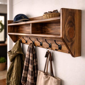 May include: A wall-mounted wooden coat rack with a top shelf and six black metal hooks. The shelf holds a small woven basket and a blue baseball cap. Hanging from the hooks are an olive green jacket, a brown plaid scarf, and a beige tote bag.
