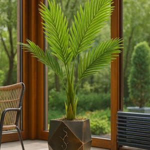 May include: A faux palm tree in a geometric bronze-colored pot. The plant has bright green fronds and a brown trunk. The pot sits on a wooden table in front of a window with a view of trees.