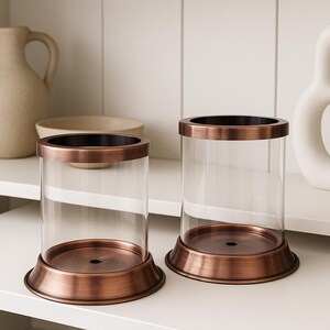 Könnte beinhalten: Zwei durchsichtige Glas-Kerzenhalter mit kupferfarbenen Metallbasen und -rändern. Die Kerzenhalter sind zylindrisch und stehen auf einem weißen Regal. Weitere dekorative Gegenstände, darunter eine Vase und eine Schüssel, befinden sich ebenfalls auf dem Regal.