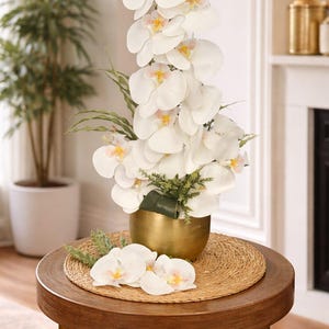 May include: A decorative arrangement of white orchid flowers with yellow and pink centers in a gold-colored metal vase. The vase sits on a round wooden table with a woven mat. Additional orchids are placed on the table.