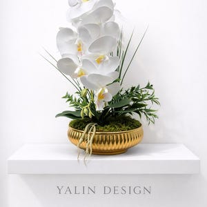 May include: A white orchid arrangement in a gold-coloured, ribbed bowl. The flowers have yellow and pink accents. The arrangement sits on a white shelf. The text "YALIN DESIGN" is below the shelf.