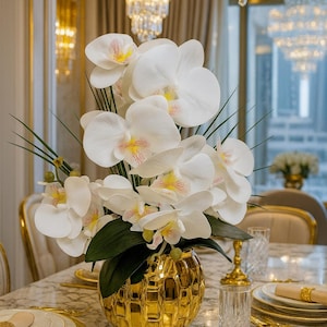 May include: A display of white orchids with yellow and pink markings, green leaves, and stems, in a gold textured vase. The arrangement is on a marble table with place settings, creating an elegant centrepiece.