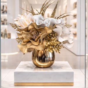 Peut inclure: Un arrangement floral décoratif dans un vase doré, composé de fleurs et de feuillage dorés, blancs et crème. Le vase est posé sur un socle en marbre blanc avec une bande dorée. L'ensemble est présenté dans une vitrine.