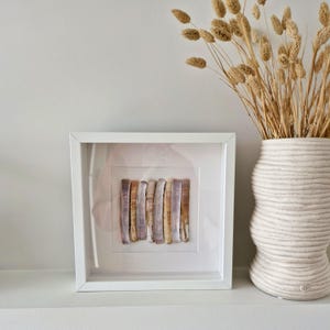 Op de afbeelding: Een ingelijst kunstwerk met een verzameling verticale, langwerpige schelpen in bruin-, beige- en paarstinten. De witte lijst staat op een wit oppervlak, met rechts een getextureerde witte vaas met gedroogd siergras.