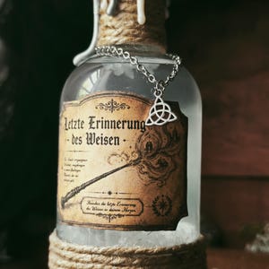 Könnte beinhalten: Eine klare Glasflasche, die an Boden und Hals mit beigefarbenem Garn umwickelt ist und weiße Wachstropfen aufweist. Eine silberne Kette mit einem Triquetra-Anhänger hängt über einem beigen Etikett mit dem Text "Letzte Erinnerung des Weisen" und einem Zauberstab.