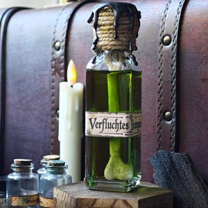 Könnte beinhalten: Eine klare Glasflasche gefüllt mit grüner Flüssigkeit und einem darin eingetauchten Knochen. Die Flasche hat ein Etikett mit dem Text Verfluchtes und einen mit beigefarbenem Garn umwickelten Verschluss, von dem schwarzes Wachs herabläuft.