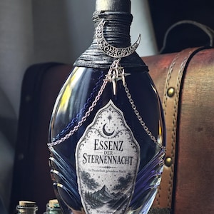 Könnte beinhalten: Eine dunkelblaue Glasflasche mit klarem Kristallverschluss und schwarzer Kordel am Flaschenhals. Ein silberner Mondsichel-Anhänger und Sternanhänger hängen an silbernen und blauen Ketten. Das Etikett trägt die Aufschrift ESSENZ DER STERNENNACHT.
