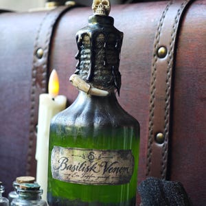 Könnte beinhalten: Eine Glasflasche gefüllt mit grüner Flüssigkeit, mit einem Etikett, auf dem Basilisk Venom und Ein Tropfen genügt steht. Der Flaschenhals ist mit Kordel umwickelt, mit einem kleinen Totenkopf verziert und mit einem weißen Fangzahn sowie schwarzem Wachs.